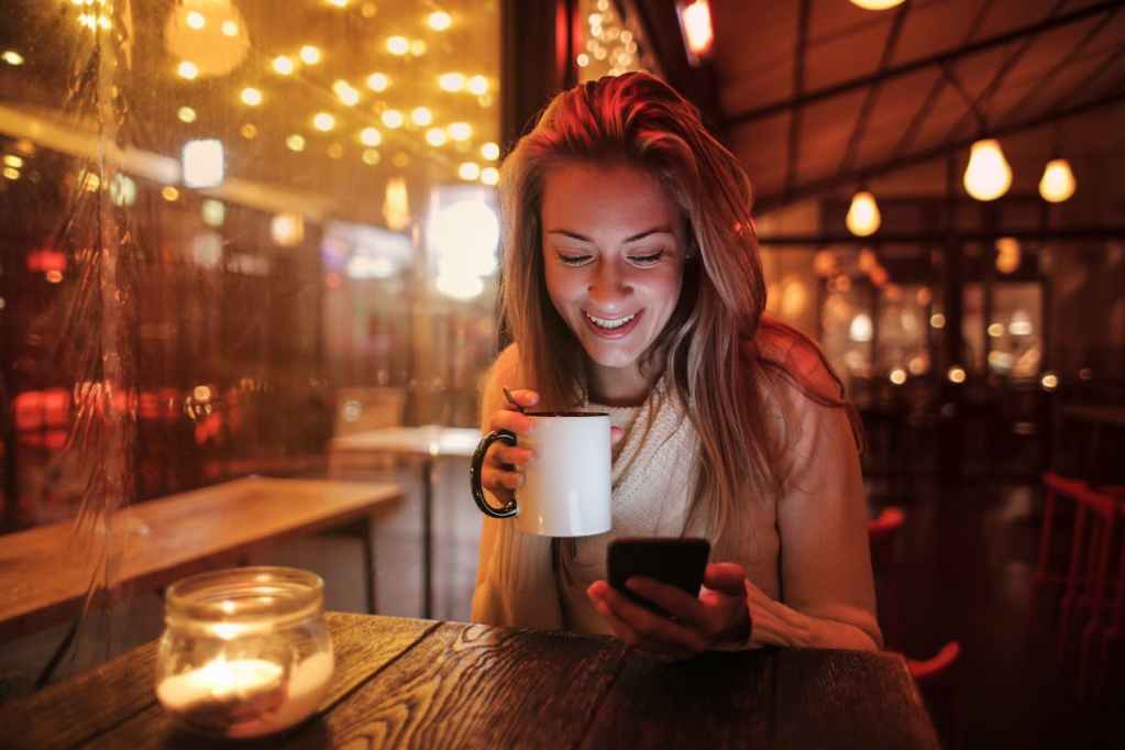 pexels photo 3783513 Woman smiling while holding coffee and using phone in warm café ambiance.