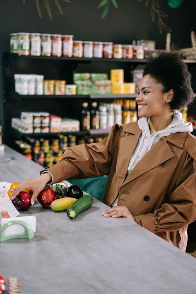 pexels photo 7990384 Woman with afro hair in a brown coat selecting fresh vegetables at a store counter, smiling.